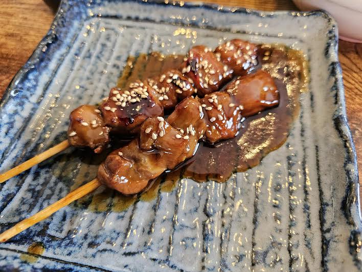 2 skewers of meat with sesame seeds and brown sauce on a square plate.